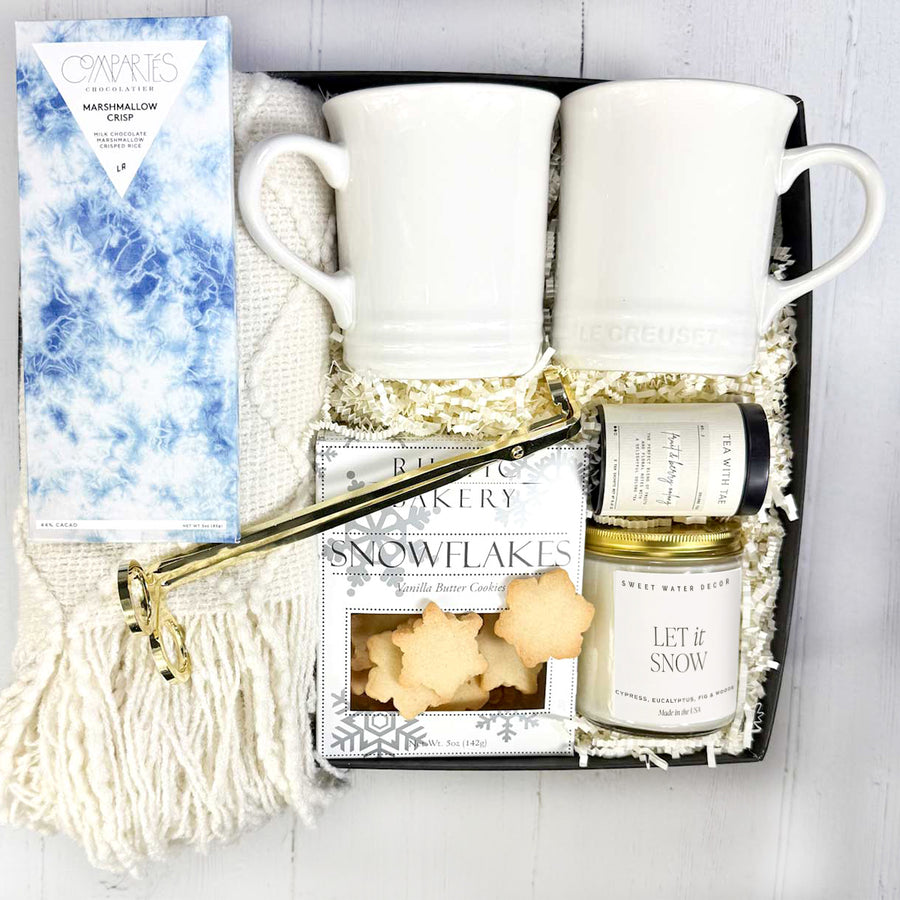 Cozy Moments Gift set with two Le Creuset mugs, a Let it snow candle, a box of snowflake cookies, tea, a throw blanket, a candle snuffer, and a gourmet chocolate bar in a box on a white background