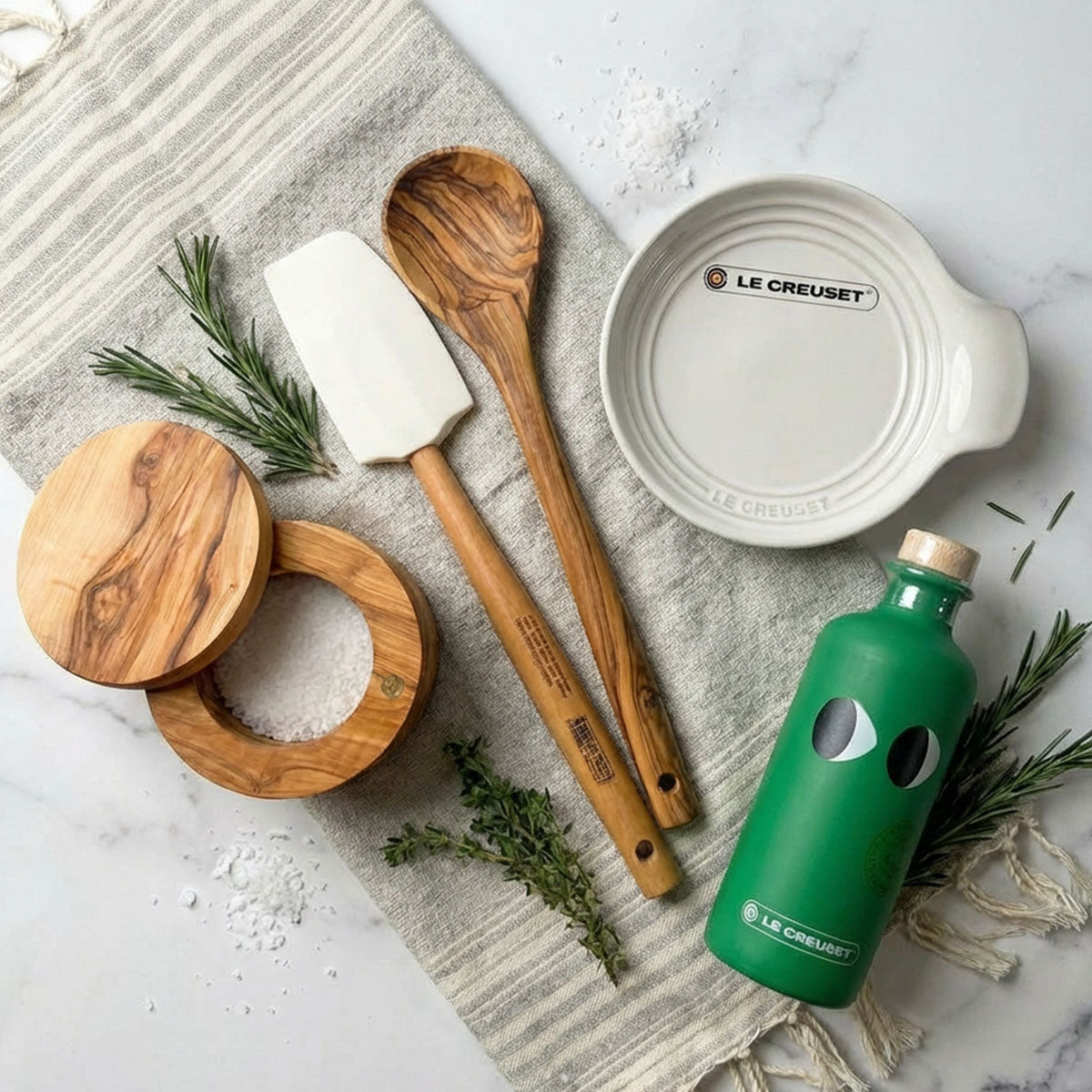Le Creuset kitchen spoon rest and spatula, wooden spoon, and olive oil on marble surface.