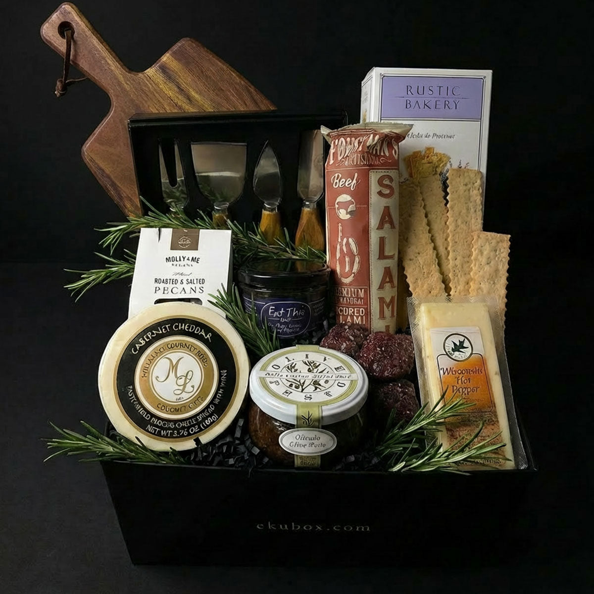 A charcuterie gift set displayed in a black box, including a wood cutting board, a 4-piece acacia wood handle utensil set, various gourmet cheeses, salami, pesto, honey, and crackers