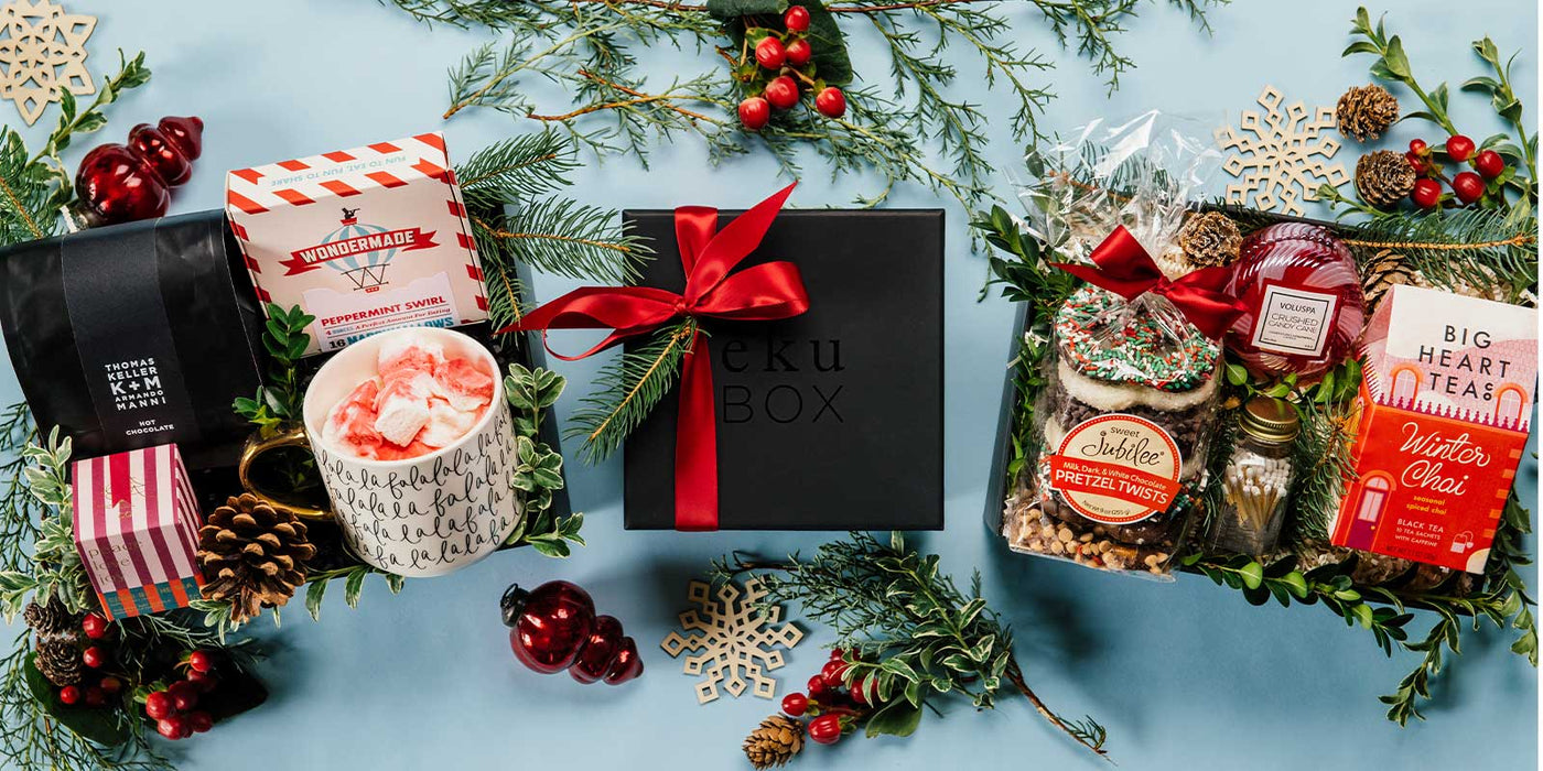 2 gift boxes with hot chocolate, tea, pretzels, a candle, matches, a mug, pine cones surrounded by holiday greens and pine cones and a ekuBOX in the middle on a light blue background/