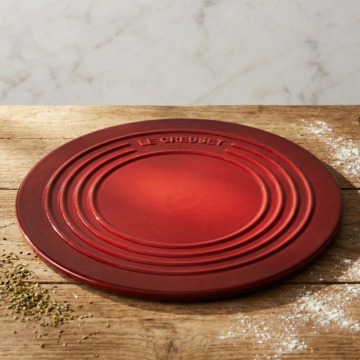 Le Creuset cerise red stoneware pizza stone on a wooden tabletop with flour and herbs.