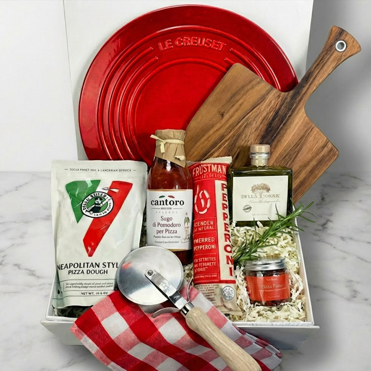 Gourmet Pizza Party box with pizza-making items including a red Le Creuset dish, pizza dough, and tools on a white surface.