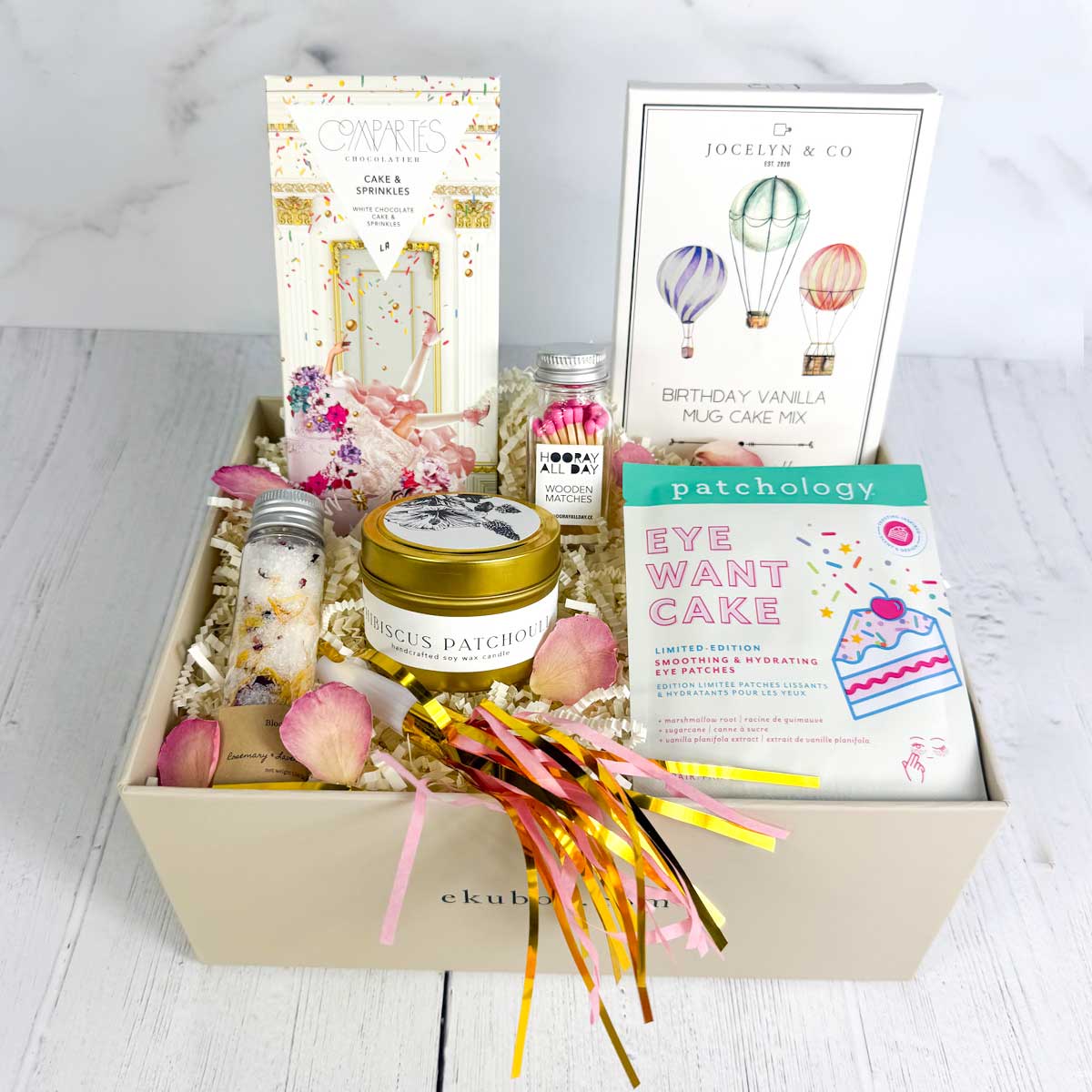 Birthday gift box featuring cake-themed treats and self-care items, including a vanilla mug cake mix, sprinkle chocolate bar, candle, colorful matches, eye patches, floral bath salts, and a party blow horn arranged in a small gift box.