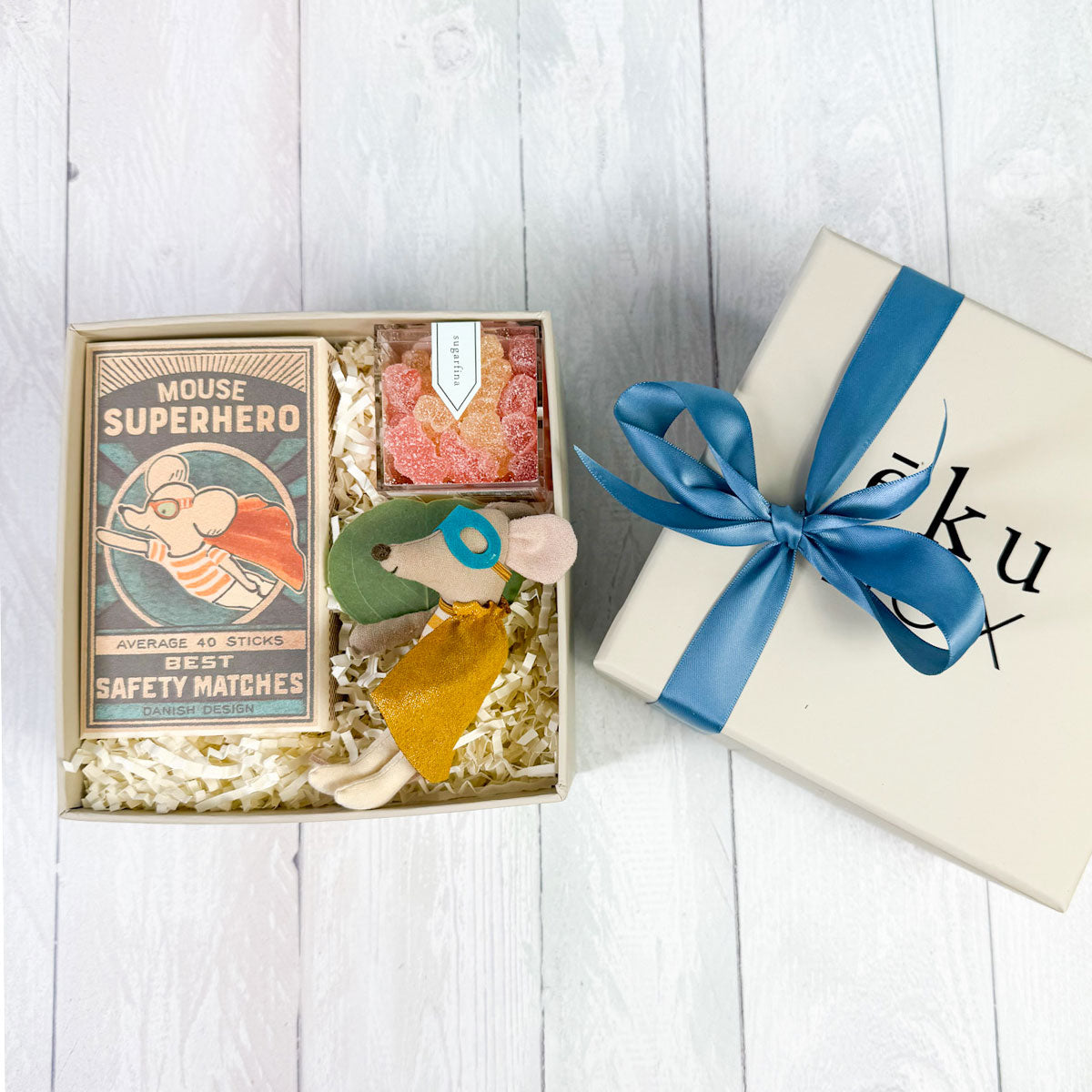 Top view of A gift box with a Maileg Superhero Mouse in a matchbox, accompanied by a plush mouse, a gummy treat, and a decorative item.