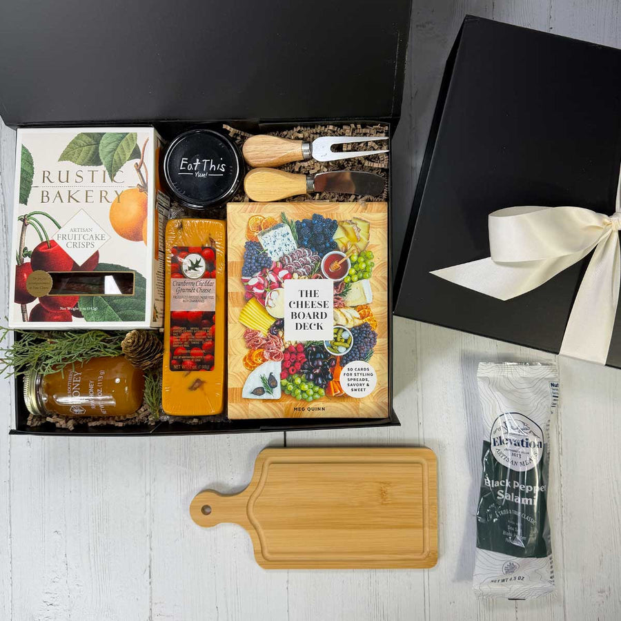 small charuterie gift box with crackers, salami, box of cheese board deck cards, cheddar cranberry cheese, a honey jar, fig jam, and utensils in a black box on a grey background