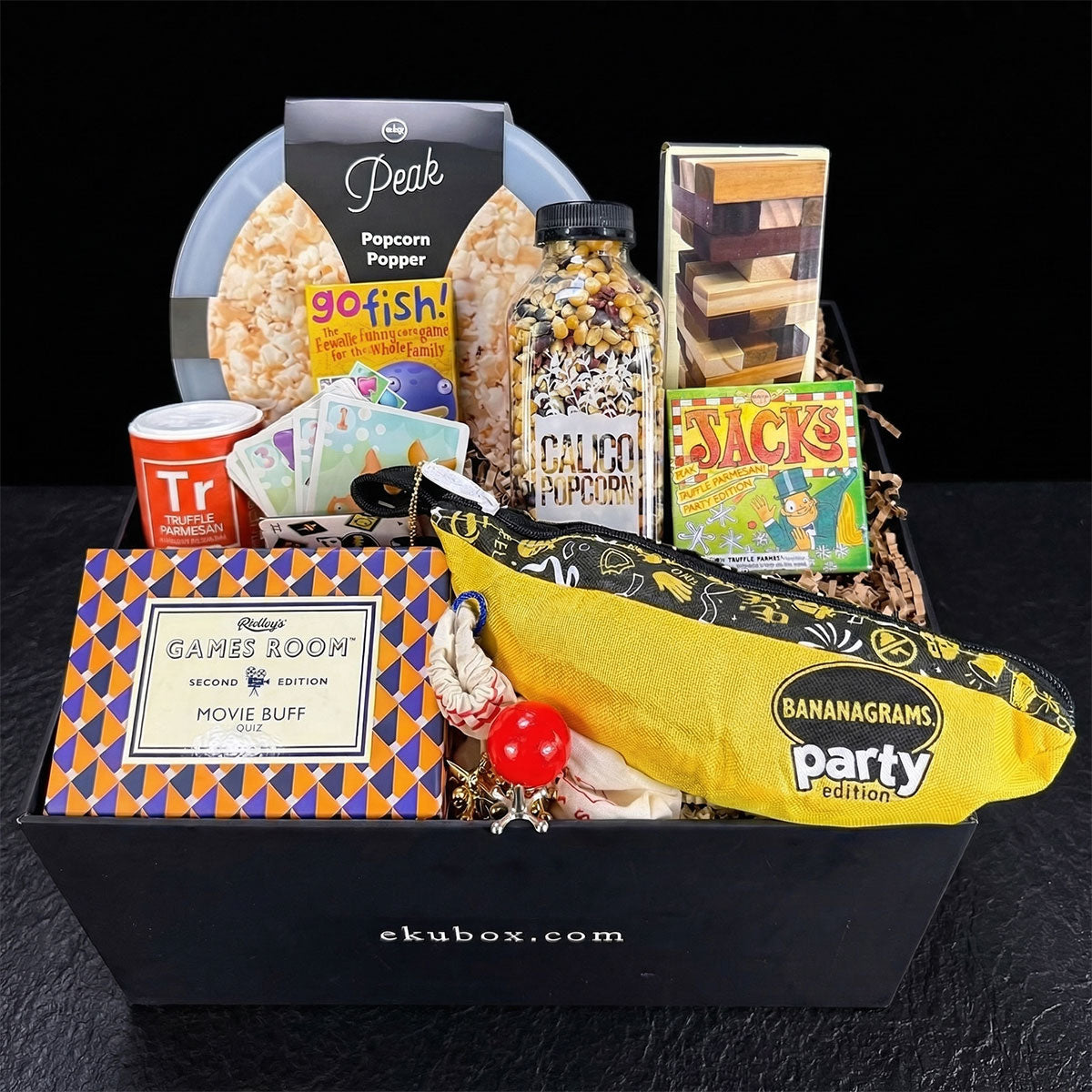 Gift box with games,  banangrams, movie buff trivia cards, jacks, snacks, popcorn kernals and a popcorn popper on a dark surface.