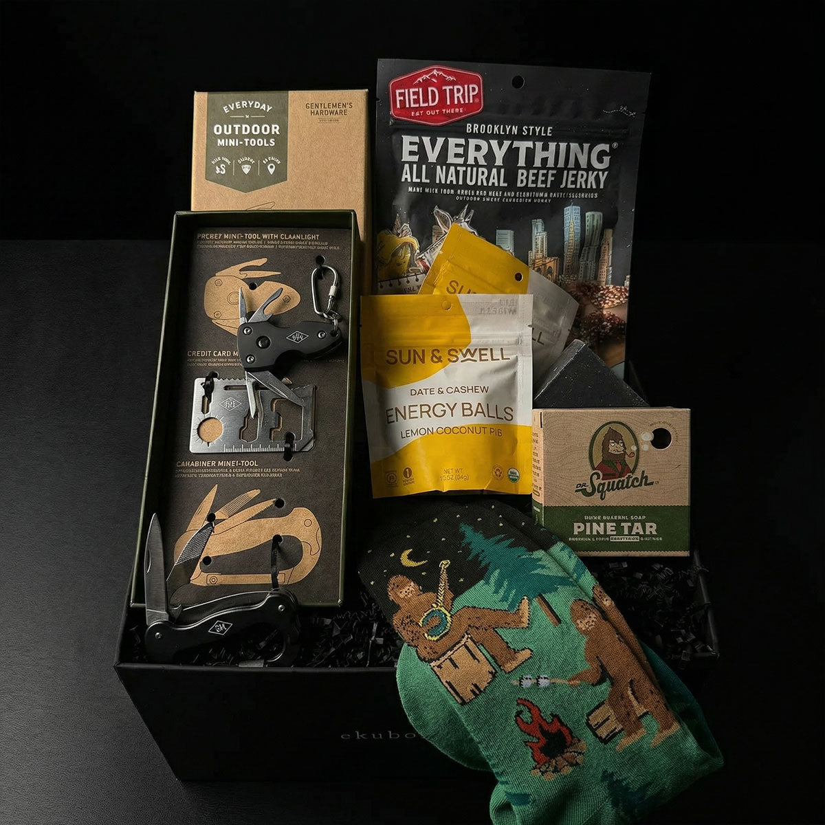 A collection of outdoor-themed items including a Sasquatch-themed sock, a pine tar bar of soap, a mini toolkit with a multi-tool with a flashlight, screwdriver, and other tools, a package of gourmet jerky