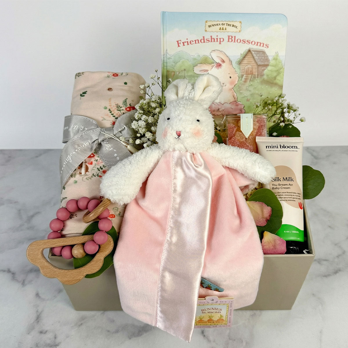 Newborn Baby girl gift box with a pink bunny lovey, floral swaddle, baby book, teether, and body cream in box on marble background