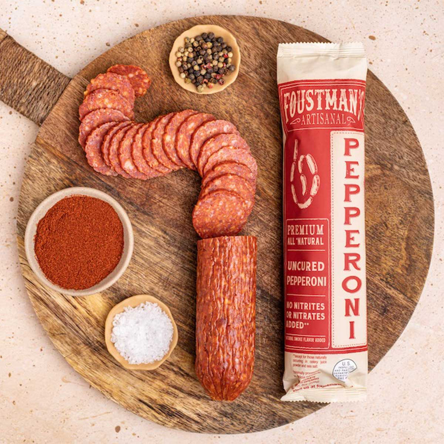 Wooden cutting board with sliced pepperoni, bowls of spices, and a Foustman Artisanal Pepperoni package.