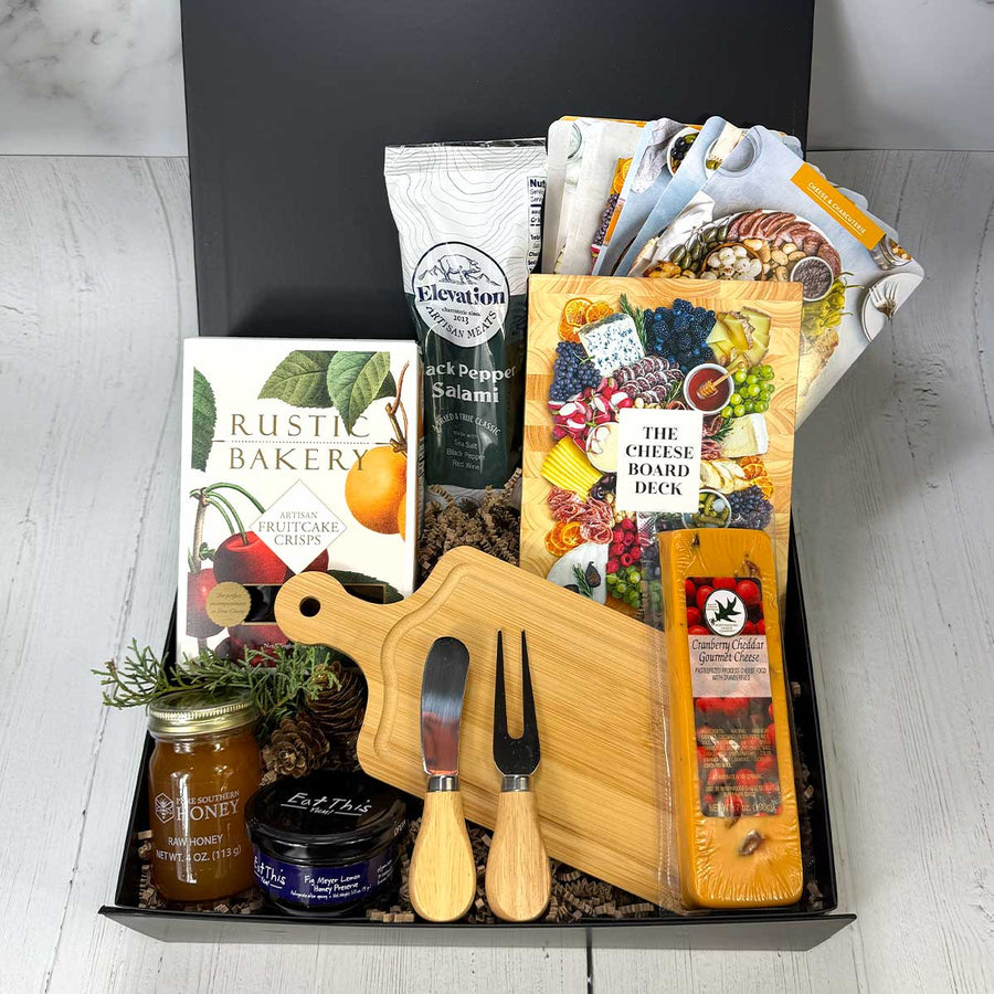 small charuterie gift box with crackers, salami, box of cheese board deck cards, cheddar cranberry cheese, a honey jar, fig jam, and utensils in a black box on a grey background