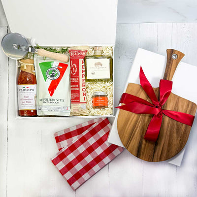 ekuBOX Gift box with pizza-making items including dough mix, pizza sauce sauce, pepperoni, olive oil, pizza seasoning, red and white checked kitchen towel and a pizza cutter on a white wood surface. Pizza paddle with a red bow on a white gift box.