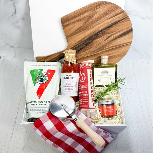 ekuBOX Gift box with pizza-making items including dough mix, pizza sauce, pepperoni, olive oil, pizza seasoning, red and white checked kitchen towel, pizza paddle, and a pizza cutter on a white wood surface. 