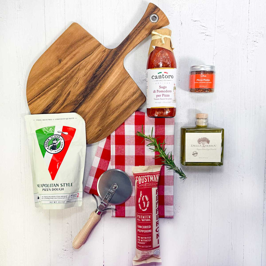  pizza-making items including dough mix, pizza sauce, pepperoni, olive oil, pizza seasoning, red and white checked kitchen towel and a pizza cutter on a white wood surface.