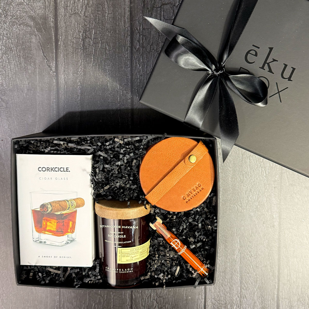 Cigar glass gift box with a Corkcicle cigar rocks glass, whiskey-infused toothpicks, tobacco flower candle, and four leather coasters on a dark surface. 