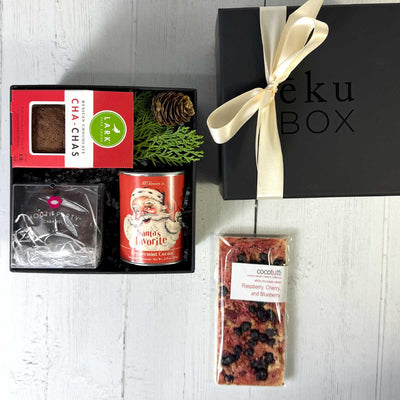Gift items including a box of 'Chachas', a can of 'Santa's Favorite' cocoa, and a gourmet white chocolate bar on a white surface with a 'eku Box' branded box.