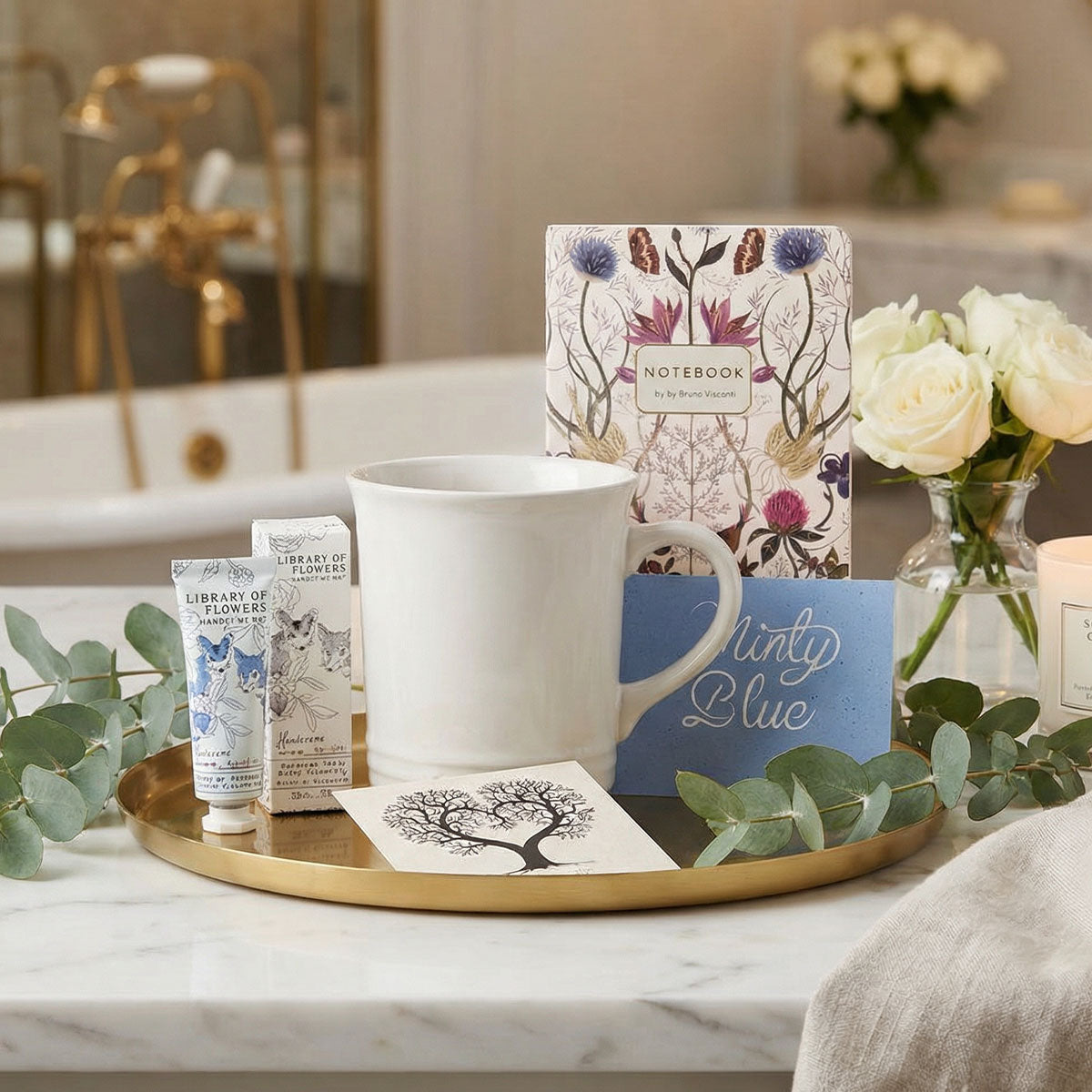 White mug on a gold tray with floral notebook, tea packet, and flowers in a bathroom setting.