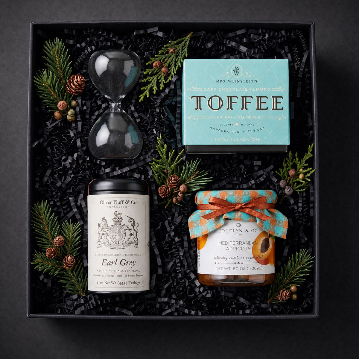 gift box with a sand timer, tin of tea, box of toffee and a jar of dried apricots with fresh winter greens
