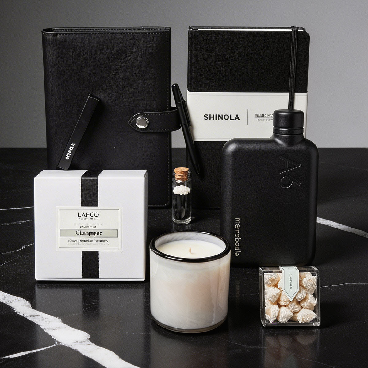 black marble surface with a Shinola notebook cover, shinola notebook, shinola stainless pen, a White candle with black rim and box, a memobottle flask, candies and matches on it.