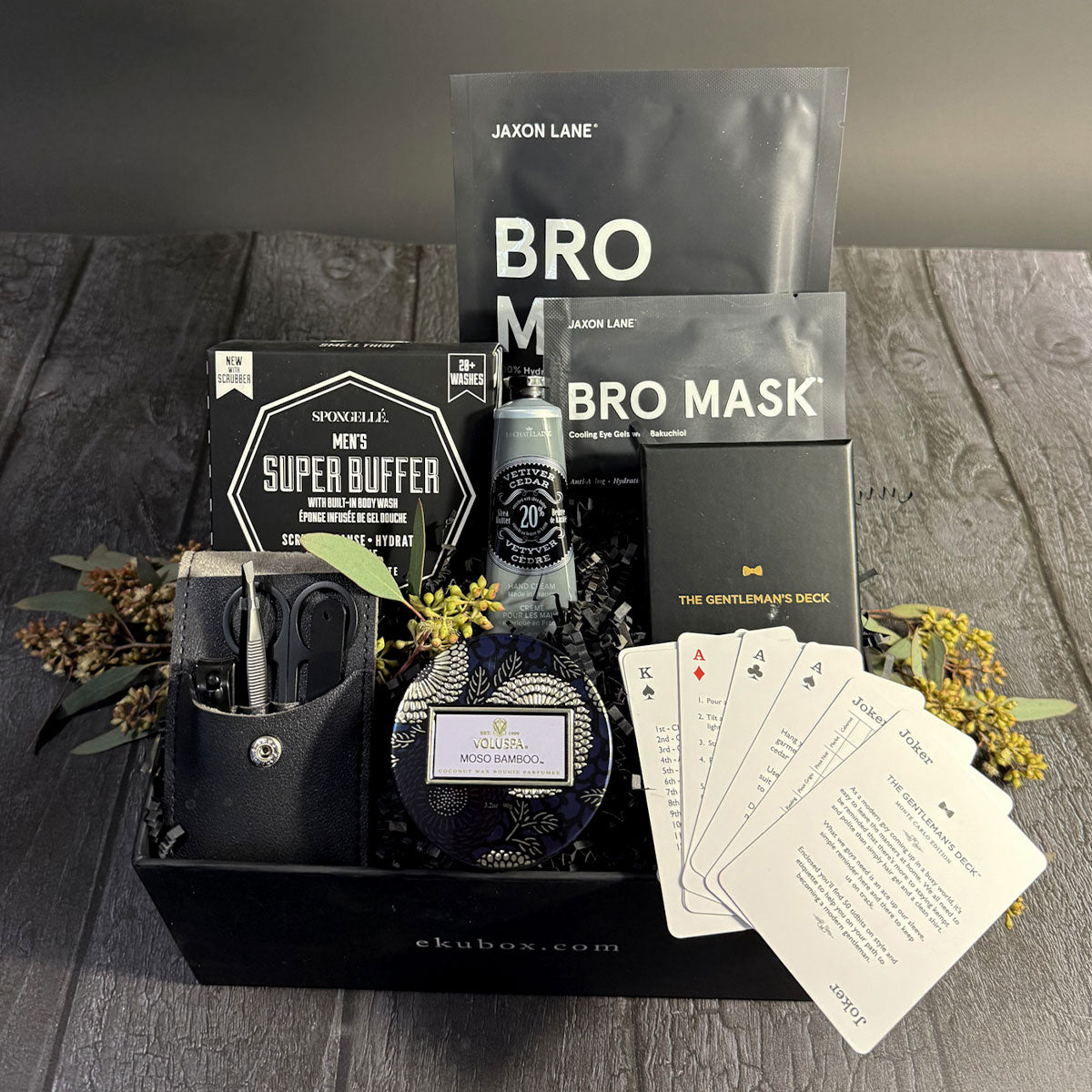 A gift box set containing men's grooming essentials including a candle, hand cream, manicure set, sheet mask, and eye patches, all displayed on a wooden surface with a black background.
