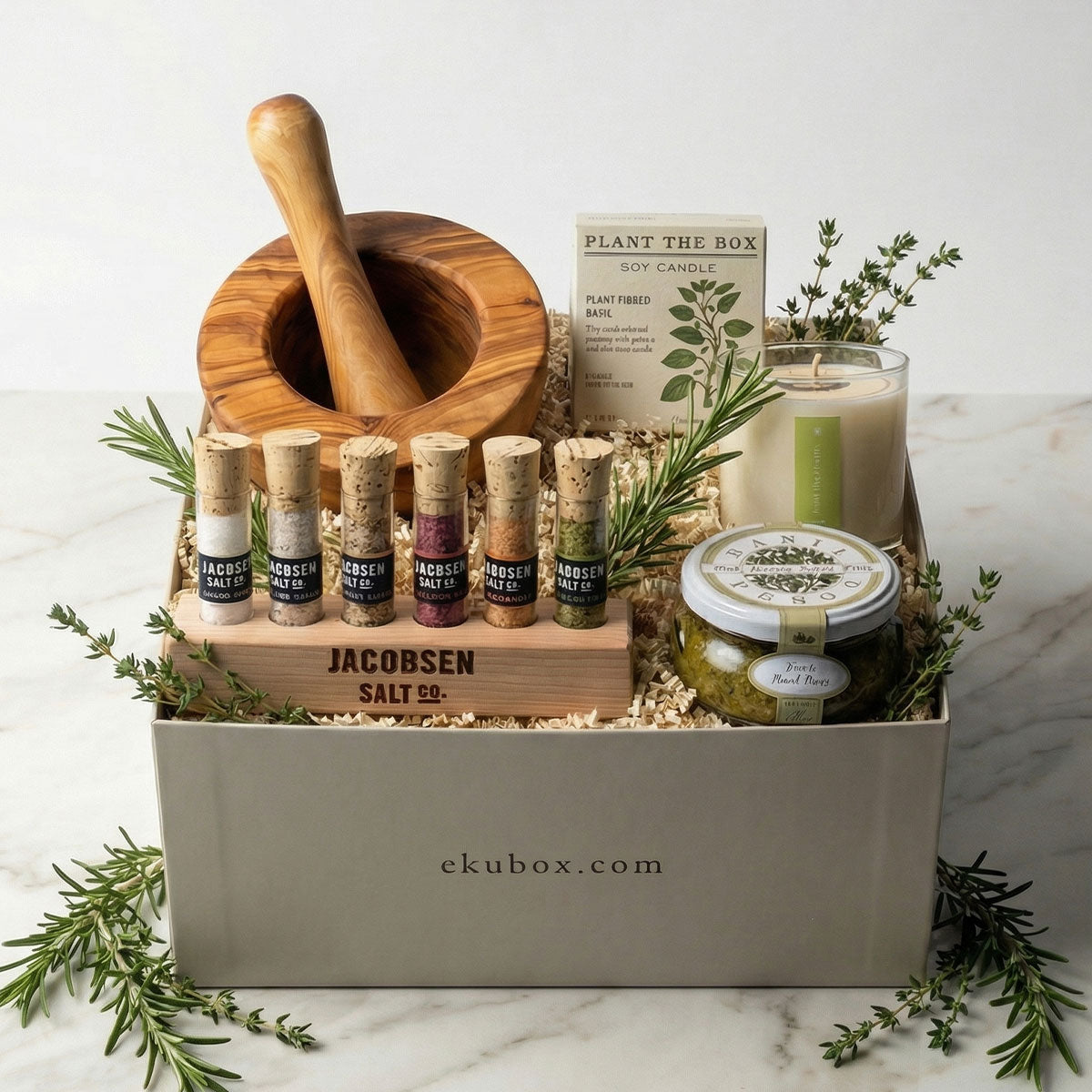 Herbalist gift box with wooden mortar and pestle, plant the box candle, and herb salt test tubes and basil pesto on a marble surface.