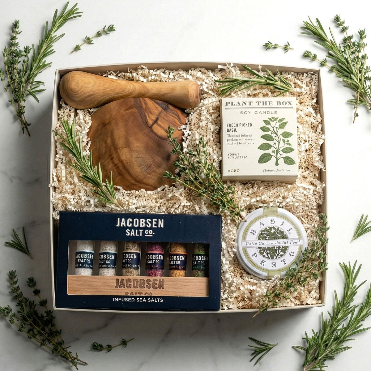 Herbalist gift box with wooden mortar and pestle, plant the box candle, and herb salt test tubes and basil pesto on a marble surface.