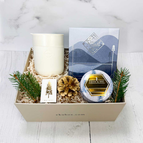 Gift box with candle, box of drinking chocolate, a cream colored ceramic mug, a box of matches and decorative elements on a white wood surface