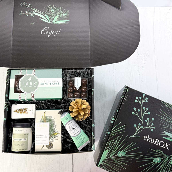 Mailer ekuBOX Gift box with various items including a tea canister, Mint chocolate cookies, sugarfina chocolate caramels, pine candle in a decorative box, seasonal matches and a handcream, on a white surface. It shows the top of the ekuBOX MAILER BOX ON THE RIGHT SIDE- A BLACK BOX WITH GREEN FLORAL DESIGN