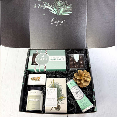 Mailer ekuBOX Gift box with various items including a tea canister, Mint chocolate cookies, sugarfina chocolate caramels, pine candle in a decorative box, seasonal matches and a handcream, on a white surface.