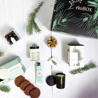 Mailer ekuBOX Gift box with various items laying flat on a white background - including a tea canister, Mint chocolate cookies, sugarfina chocolate caramels, pine candle in a decorative box, seasonal matches and a handcream.